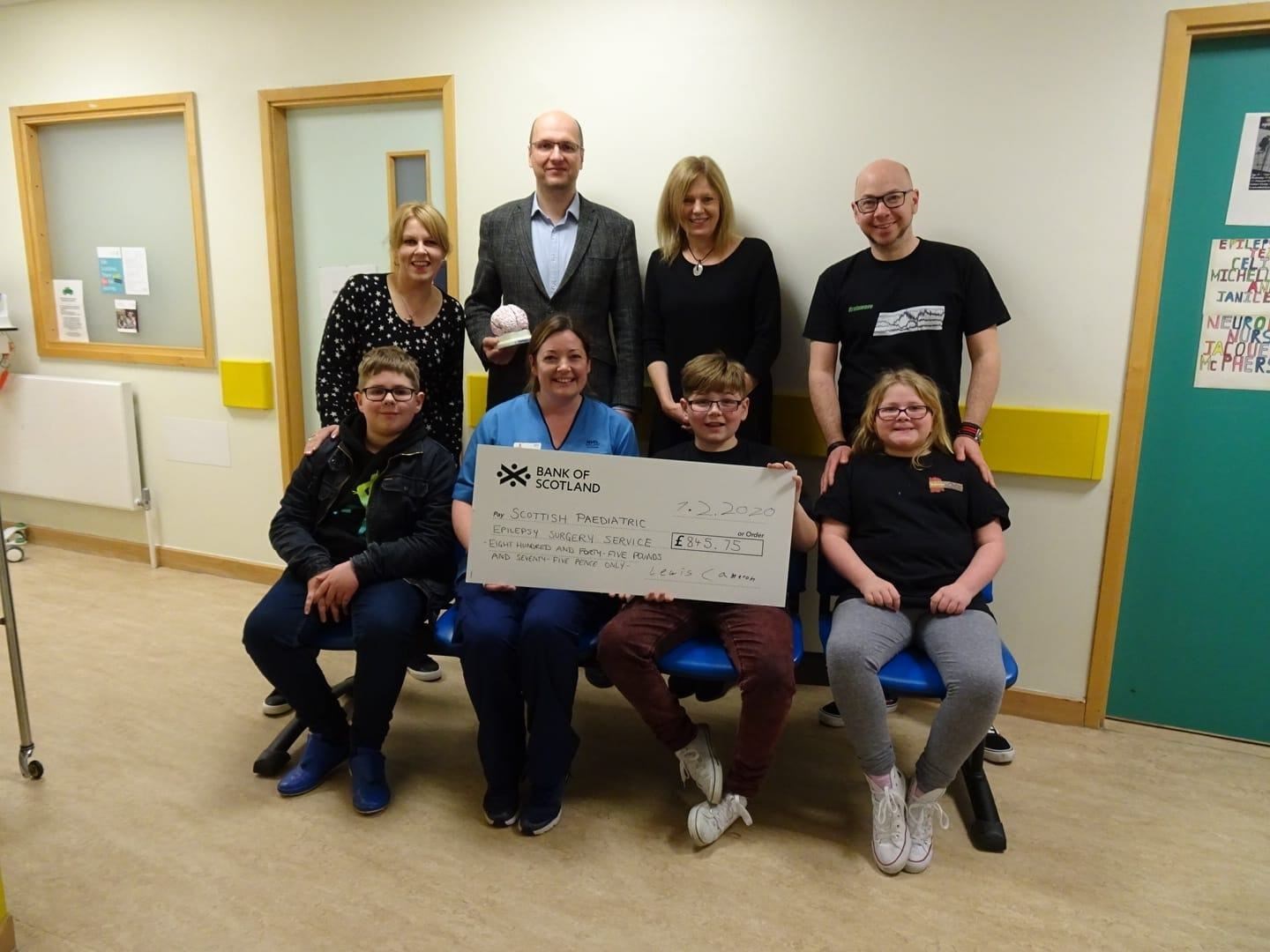 Lewis (front second right) hands over his cheque to Epilepsy surgery nurse Janice Fyall. Joining him is brother Fraser, sister Pearl and (back from left) mum Sarah Roger, neurosurgery specialist Drahoslav Sokol, Dr Ailsa McLellan and dad Garry Cameron.