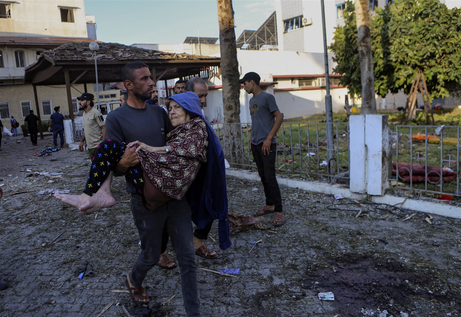 A Palestinian man carries an elderly woman past the site of a deadly explosion at al Ahli hospital (Abed Khaled/AP)