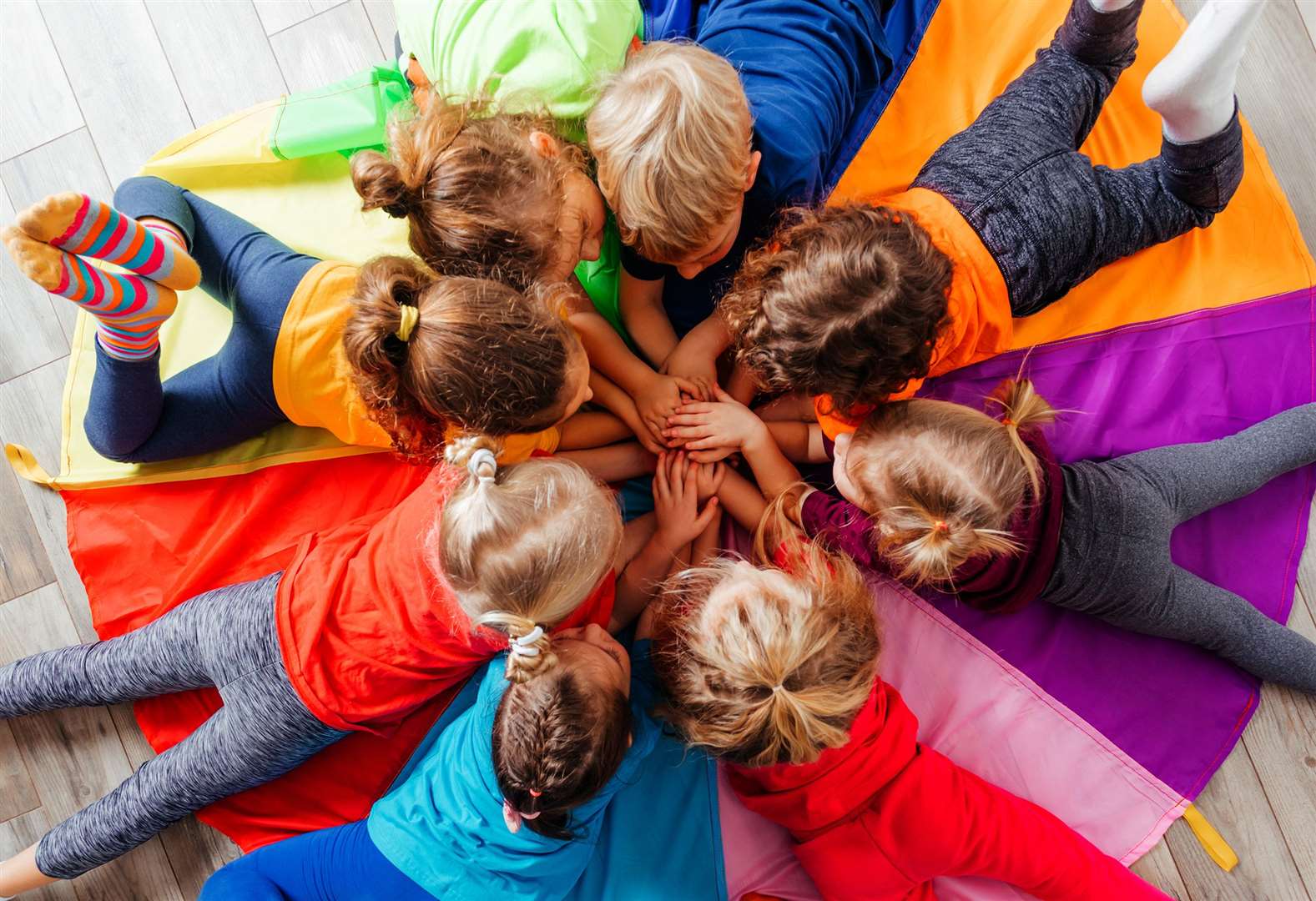Lovely children laying on multicolor canopy in circle. Team building game for daycare children in colorful t-shirts. Top view kids on wooden floor.